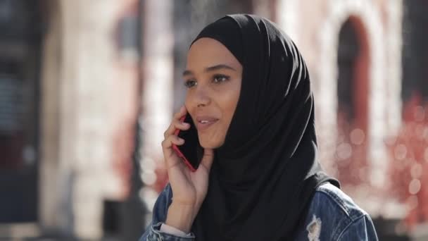 Sonriente joven musulmana que usa pañuelo para la cabeza hijab hablando con amigos en un teléfono inteligente en la ciudad, una dama camina por la vieja calle en negro hijab . — Vídeo de stock