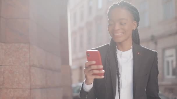 Elegante mujer de negocios afro utilizando smartphone caminando por la calle cerca del centro de negocios. Negro con estilo. Ricitos. Peinado afro . — Vídeos de Stock