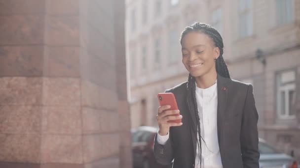 Elegante mujer de negocios afro utilizando smartphone caminando por la calle cerca del centro de negocios. Negro con estilo. Ricitos. Peinado afro . — Vídeos de Stock