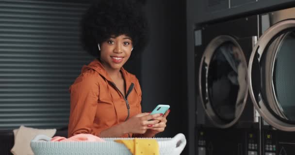 Retrato de la joven mujer afroamericana feliz con teléfono inteligente mirando a la cámara. Fondo de lavandería pública de autoservicio. Primer plano . — Vídeo de stock