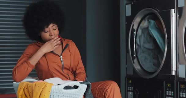 Tired young African American woman reading a book while washing her laundry at laundromat. She yawns and waits for the completion of the process. — Stock Video