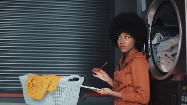 Young attractive African American women writing "to do list" on notepad sitting in the self-service public laundry. — Stock Video