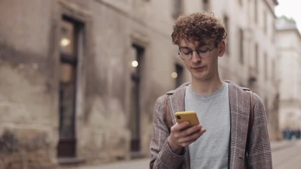 Joven chico hipster con gafas charlando con amigos en las redes sociales y escuchar música, utilizando un dispositivo de teléfono inteligente moderno mientras camina por la calle de la ciudad. Estilo de vida urbano . — Vídeo de stock