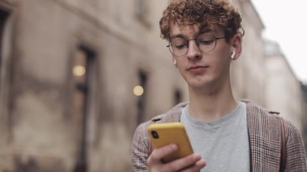 Jeune hipster avec des lunettes bavarder avec des amis sur les réseaux sociaux et écouter de la musique, en utilisant un smartphone moderne tout en marchant dans la rue de la ville. Mode de vie urbain . — Video