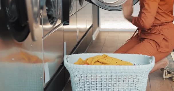Genç Afro-Amerikalı kadın bir çamaşır makinesi önünde oturur ve kirli çamaşır ile yıkayatıcı yükler. Self servis kamu çamaşırhanesi. — Stok video