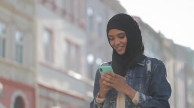 Genç gülümseyen Müslüman kadın şehir merkezinde hijab başörtüsü ayakta giyen ve akıllı telefon kullanarak. İletişim, online alışveriş, sosyal ağ konsepti.