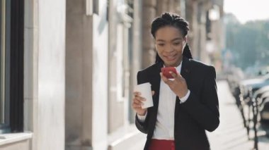 Bir takım elbise Kadın genç Afrikalı Amerikalı iş kadını bir yaz günü bir şehir caddesinde yürüyüş kahve içme online bir akıllı telefon ses tanıma fonksiyonu kullanarak.