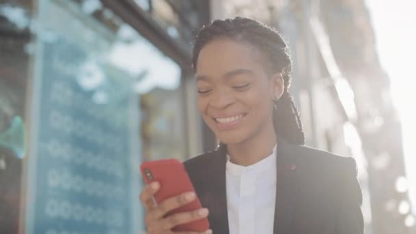 Feliz mujer de negocios afro utilizando teléfono inteligente de pie en la calle cerca del centro de negocios. Negro con estilo. Ricitos. Profesional femenino de traje que tiene buenas noticias en el teléfono inteligente . — Vídeo de stock