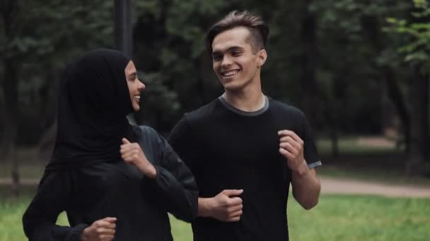 Junge kaukasische Mann und Musselin Frauen tragen Hijab schauen aufgeregt und entspannt lächelnd und plaudernd im Park Seitenansicht Fokus auf den Mann. — Stockvideo