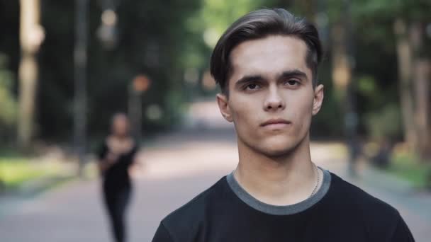 Retrato de un joven guapo parado en el parque y una joven corriendo sobre el fondo . — Vídeo de stock