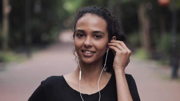 Retrato da jovem mulher bonita usando fones de ouvido de pé no parque Close Up. Conceito de estilo de vida saudável . — Vídeo de Stock