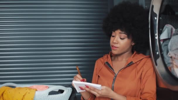 Unga attraktiva afroamerikanska kvinnor skriver "att göra-lista" på antecknings block sitter i självbetjäning offentlig tvätt. — Stockvideo