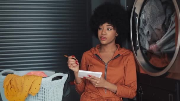 ผู้หญิงแอฟริกันอเมริกันสาวที่น่าสนใจ เขียน "ทํารายการ" บนแผ่นบันทึก นั่งอยู่ในห้องซักรีดสาธารณะ . — วีดีโอสต็อก