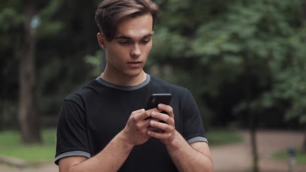 Красивый молодой человек, выглядящий изумленным, прокручивая телефон и говоря: Вау, стоя в парке . — стоковое видео