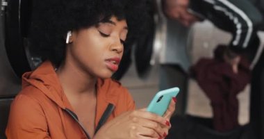Genç African American kadın Laundromat akıllı telefon kullanarak gülümseyen. Müzik dinliyor ve şarkı söylüyor. Self-Servis kamu Çamaşırhane.
