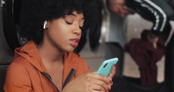 Jeune femme afro-américaine souriante utilisant un smartphone à la laverie automatique. Elle écoute de la musique et chante. Laverie publique en libre-service . — Video