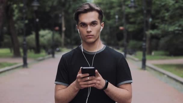 Portrait d'un jeune homme aspirant dans un casque et d'un téléphone intelligent dans sa main debout dans un parc Concept Mode de vie sain  . — Video