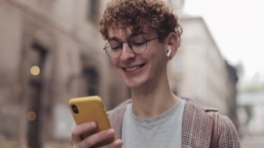 Yuvarlak Gözlük ve Kulaklık genç gülümseyen Millennial Eski Şehir Caddesi Yürürken Onun Modern Smartphone kullanma. İletişim, Öğrenci, Turist Kavramı. Kentsel Yaşam Tarzı.