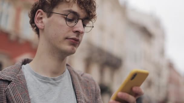 Close Up Portrait of Serious Millennial in Glasses Usando seu aplicativo de celular moderno, Olhando para a tela, Bebendo café, em Blured City Background. Vista lateral . — Vídeo de Stock
