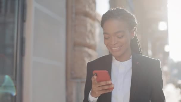 Happy afro affärskvinna med smartphone stående på gatan nära Business Center. Svart snygg. Dreadlocks. Kvinnlig professionell i kostym ha goda nyheter på smartphone. — Stockvideo