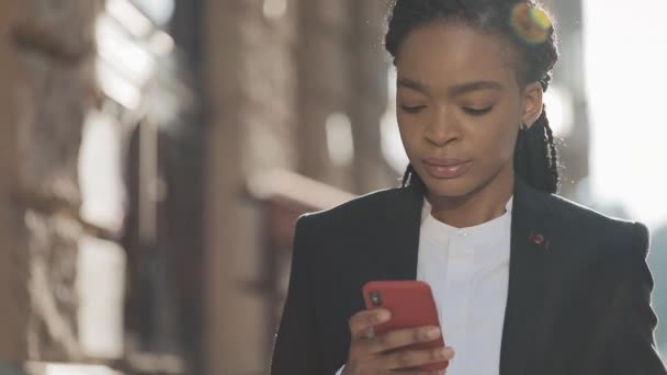 Empresaria cansada usando smartphone caminando por la calle cerca del centro de negocios. Negro con estilo. Ricitos . — Vídeo de stock