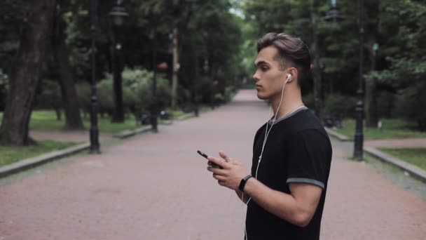 Portrair of Handsome Young Man in Headphones and Smartphone in his Hand Wearing Fitness Tracker Standing in Park and Turning his Head Concept Healthy Lifestyle. — Stock Video