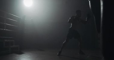 Siluet boksör adam karanlık spor salonunda kum torbası vurur. Dövüşçü egzersizi. Erkek boksör karanlıkta egzersiz. Kickboxer siluet eğitimi.