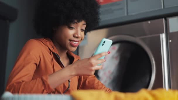 Mujer joven sonriente usando teléfono inteligente en la lavandería. Autoservicio de lavandería pública . — Vídeo de stock