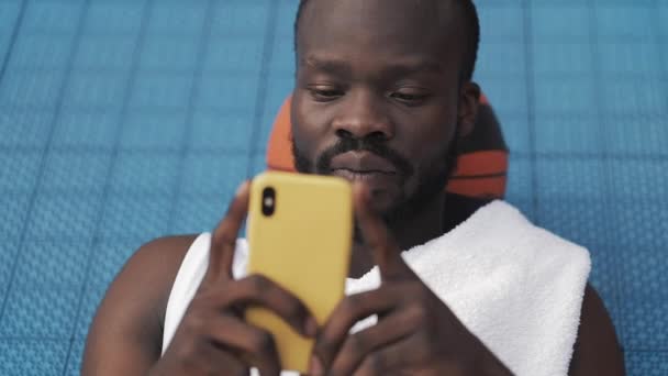 Bliska Afro American Guy leżącego na ziemi z piłką pod głową, za pomocą swojego smartfona do rozmów w Urban Sports koszykówki sądu. Zdrowy styl życia i Sport Concept. Widok z przodu. — Wideo stockowe