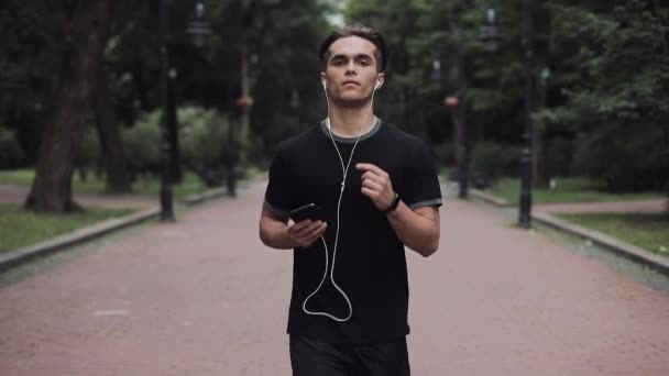 Joven guapo en auriculares y Smartphone en su mano corriendo en el concepto de parque estilo de vida saludable . — Vídeo de stock