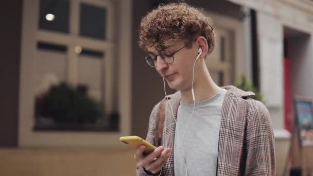 チェックジャケットを着たヘッドフォンでメガネの若いヒップスター赤いカーリーヘアガイのクローズアップ。彼のスマートフォンを使用して、ウィットの友人をチャット。旧市街の背景を歩く。学生,観光コンセプト. — ストック動画