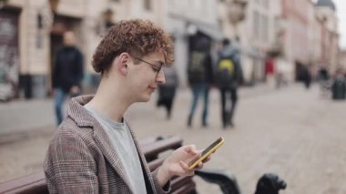 Genç Hipster Guy'ın Gözlük ve Ceketle Yan Görünümü Akıllı Telefonunu Kullanarak, Ekranda Kaydırma, Eski Şehir Arka Plan Close Up'ta Bankta Otururken Gülümseme.