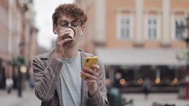 Gözlüklü ve Kulaklıklı Genç Hipster Guy, Sosyal Ağlarda Akıllı Telefon Yazma Mesajını Kullanarak Kontrol Edilmiş Ceket Giyiyor, Old City Caddesi'nde Kahve İçmesi. İletişim Kavramı.