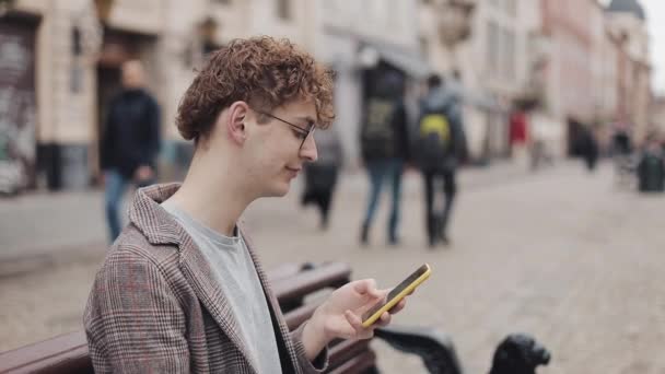 Seitenansicht eines jungen Hipsters in Brille und Jacke, der sein Smartphone benutzt, auf dem Bildschirm blättert und lächelt, während er auf der Bank vor Altstadtkulisse sitzt. — Stockvideo