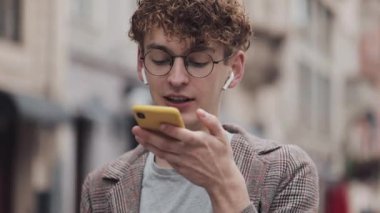 Gözlüklü ve Kulaklıklı Genç Hipster Guy Onun Smartphone Ses Komutanı kullanarak Ceket Giyen veya Sesli Mesaj Yapma, City Street Ayakta.