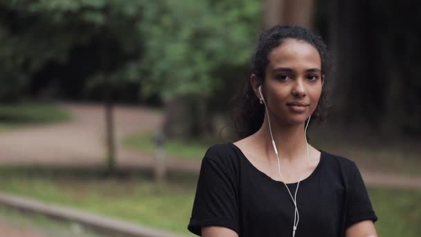 Retrato de bastante joven Musline Girl en auriculares de pie en el parque girando la cabeza y sonriendo concepto de estilo de vida saludable . — Vídeos de Stock