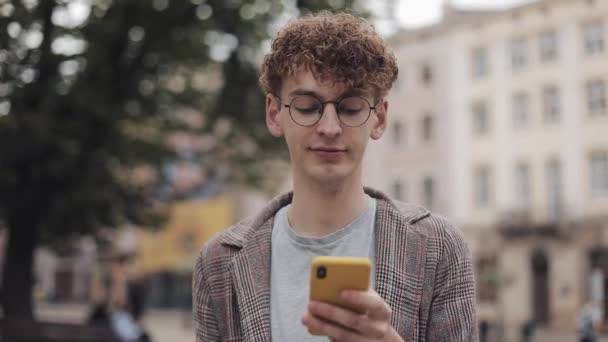 Primo piano del ragazzo giovane studente hipster in occhiali e auricolari wireless che indossa giacca utilizzando il suo telefono cellulare, sorridente, bere caffè mentre cammina a Old City Street . — Video Stock