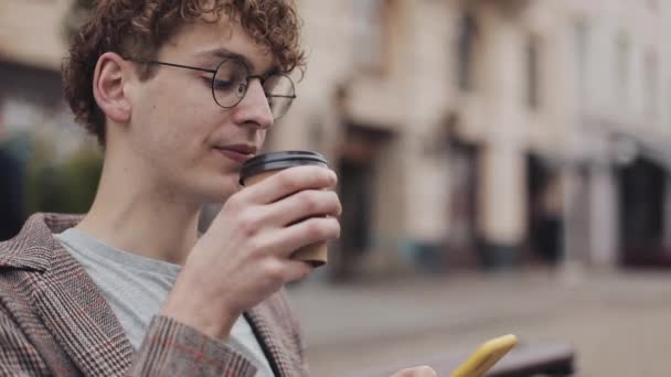 ジャケットとメガネを着用した学生の男がコーヒーを飲み、シティストリートサイドビューのベンチに座っている間、彼の近代的な携帯電話の画面をスクルーリングする学生の男のクローズアップ. — ストック動画