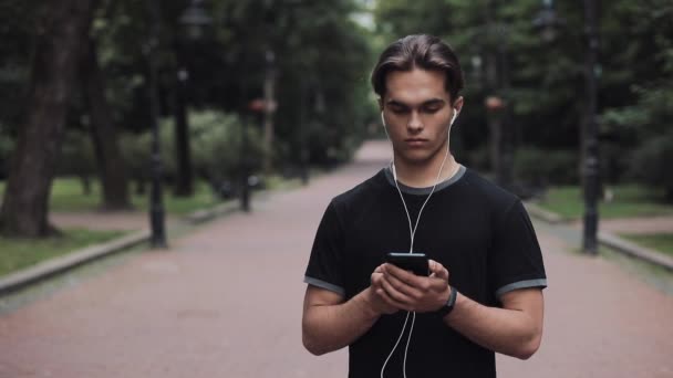 Joven guapo con auriculares y Smartphone en la mano usándolo de pie en el parque Slow Motion Concept Vida Saludable . — Vídeo de stock