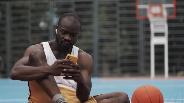 Nahaufnahme eines jungen muskulösen Afroamerikaners mit Kopfhörern, der auf dem Boden sitzt, sein Smartphone benutzt, gute Nachrichten hat und sich auf dem Street-Basketballfeld freut. Sportkonzept. — Stockvideo