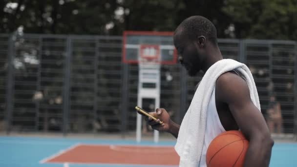 Jonge muscly serieuze Afro-Amerikaanse kerel in wit singlet Holding bal en het gebruik van zijn smartphone en het type bericht lopen op straat basketbal Hof. Technologie en sport concept. Zijaanzicht. — Stockvideo
