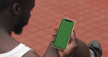 Genç Sakallı Afro Amerikan Adam Onun Smartphone kullanarak Arka Yan Görünümü, Basketbol Spor Sahası'nda Otururken Ekran dokunmadan. Spor ve Teknoloji Kavramı. Yeşil Ekran.