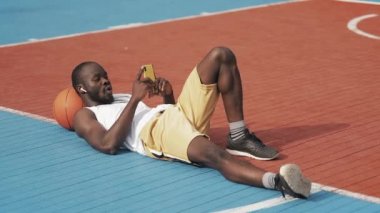 Kablosuz Kulaklıkgenç Afro Amerikan Guy Başının altında Ball ile Yere Yatan, Street Basketball Court onun Smartphone üzerinden Video arama sahip. Sağlıklı Yaşam Tarzı ve Spor Kavramı.