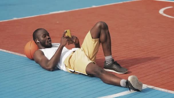 Jonge Afro Amerikaanse kerel in draadloze koptelefoon liggend op de grond met bal onder zijn hoofd, het hebben van video-oproep via zijn smartphone op straat basketbalveld. Gezonde lifestyle en sport concept. — Stockvideo
