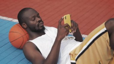 Genç Musculy Yakışıklı Afro Amerikan Guy Başının altında Topu ile yere yatarken, onun Smartphone kullanarak, Kentsel Spor Basketbol Mahkemesi'nde yazma. Sağlıklı Yaşam Tarzı ve Spor Kavramı.