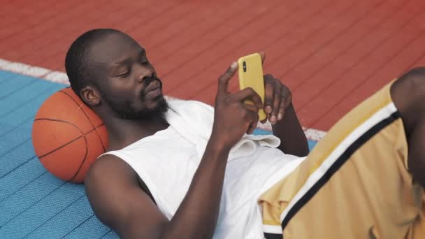 Jonge Gespierige knappe Afro Amerikaanse kerel liggend op de grond met bal onder zijn hoofd, met behulp van zijn smartphone, typen bij de stedelijke sport basketbalveld. Gezonde lifestyle en sport concept. — Stockvideo