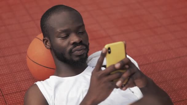 Close Up of Afro American Guy tumbado en el suelo con Ball bajo su cabeza, usando su Smartphone para escribir un mensaje en la cancha de baloncesto de deportes urbanos. Concepto de estilo de vida saludable y deporte . — Vídeos de Stock