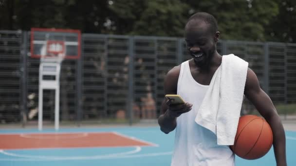 Ung muscly afro-amerikansk kille i vitt singlet hålla bollen och titta i sin smartphone, leende medan du går på gatan Basketbal domstol. Teknik-och Sportkoncept. Vy framifrån. — Stockvideo