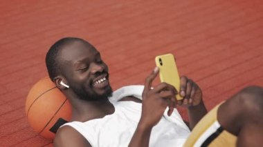 Kulaklıklı Afro Amerikalı Adam Başının Altında Topla Yerde Uzanıyor, Akıllı Telefonunu kullanıyor ve Basketbol Sahasında Gülümsüyor. Sağlıklı Yaşam Tarzı ve Spor Konsepti.