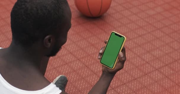 Back Side View of Afro American Man in White Singlet Χρησιμοποιώντας το Smartphone του, έχοντας μια συνομιλία μέσω βιντεοκλήσης, ενώ κάθεται στο Μπάσκετ Sport Ground. Έννοια Αθλητισμού και Τεχνολογίας. Πράσινη οθόνη. — Αρχείο Βίντεο